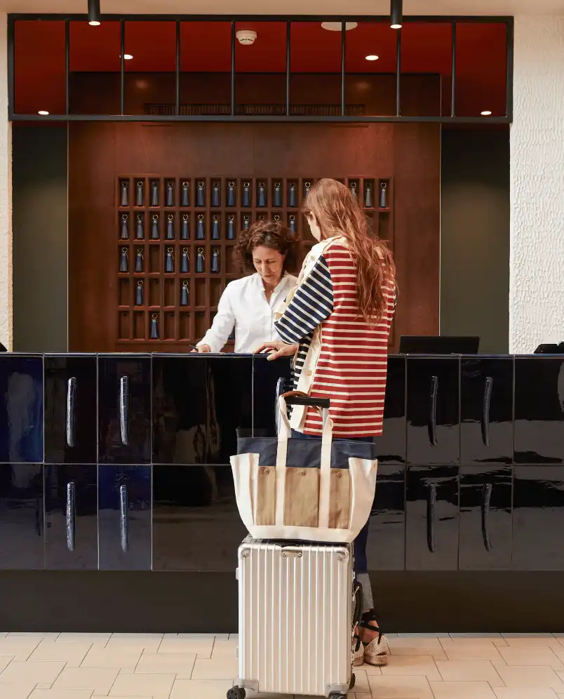 Woman with striped sweater and suitcase checking in at a hotel reception desk with a receptionist in a white shirt.