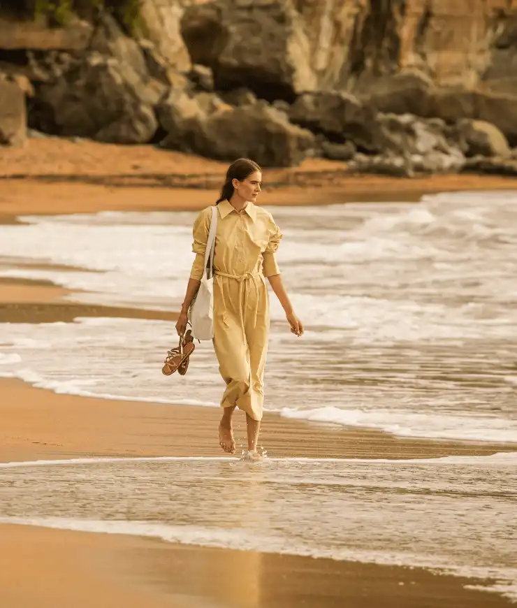 Woman walking barefoot on a sandy beach near gentle waves, holding sandals and a white tote bag, with rocky cliffs in the background.