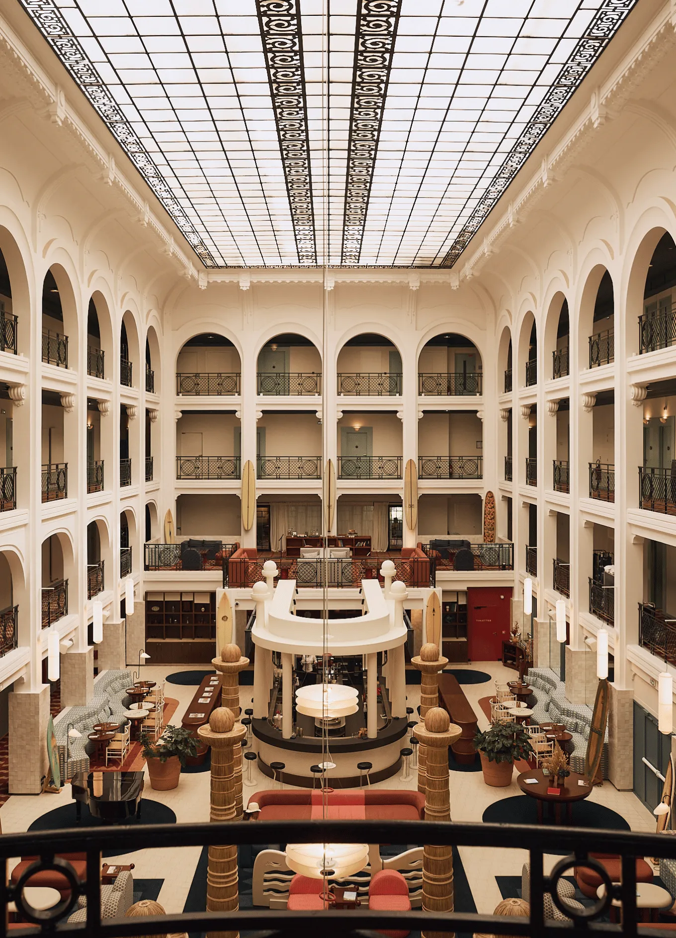 Spacious hotel lobby with large arched windows, white walls, sofas, chairs, plants, and a central reception desk under a glass ceiling.