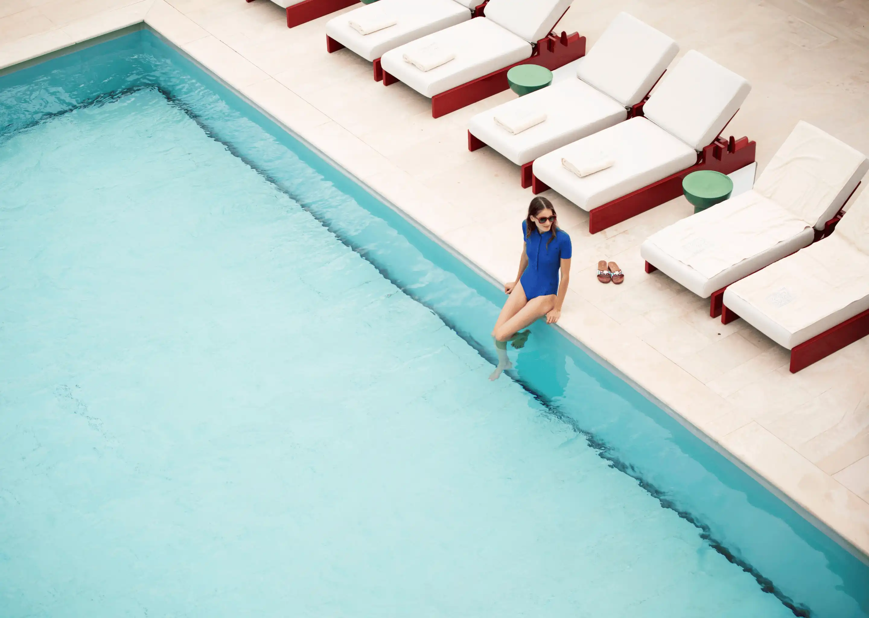 Woman in blue swimsuit with sunglasses sitting at the edge of a pool with several white lounge chairs nearby.
