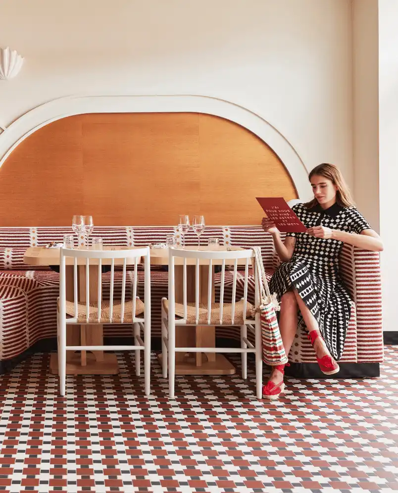Woman in a black and white patterned dress sitting on a striped bench in a restaurant, reading a menu, with patterned tiled floor and wooden wall panel behind.