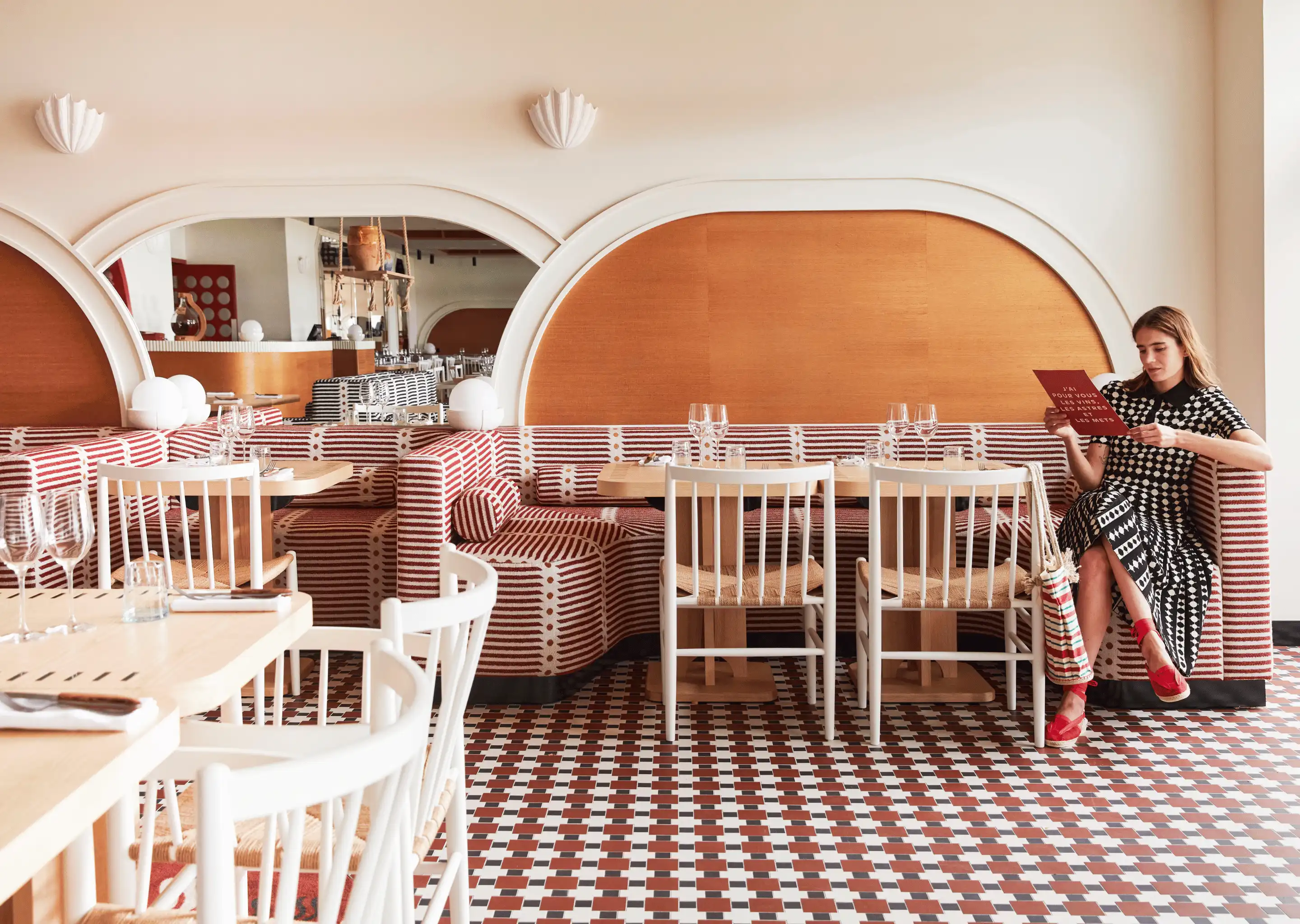 Woman in patterned dress sitting on striped red and white banquette seating in a modern restaurant with tiled floor and arched mirrors.
