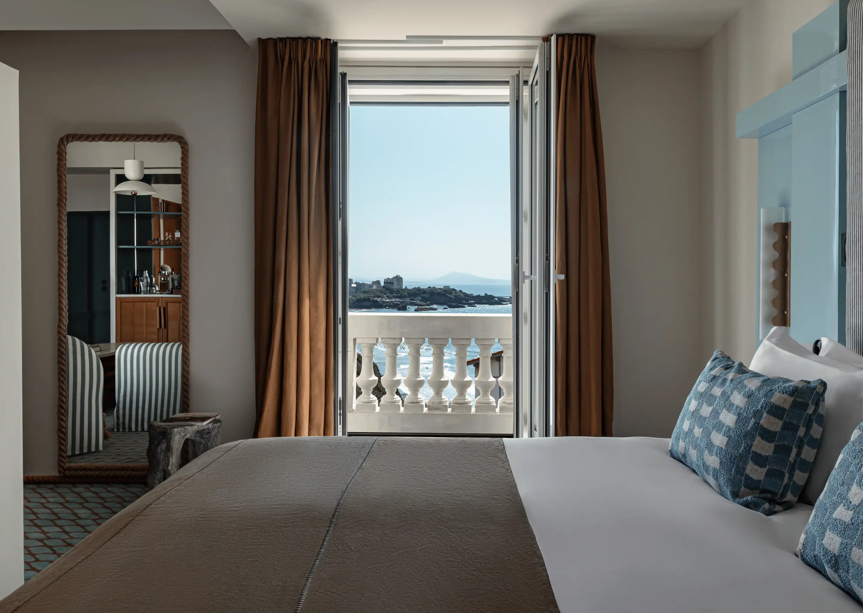 Hotel room with a bed featuring blue checkered pillows, brown curtains, an open door overlooking a balcony with a sea view.