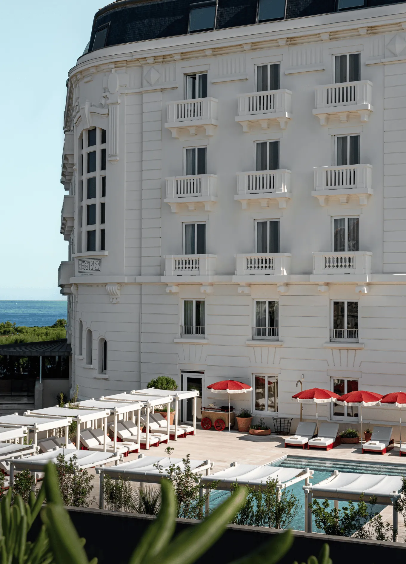 White hotel building with multiple balconies next to a pool surrounded by white lounge chairs and red umbrellas, with a view of the sea in the background.