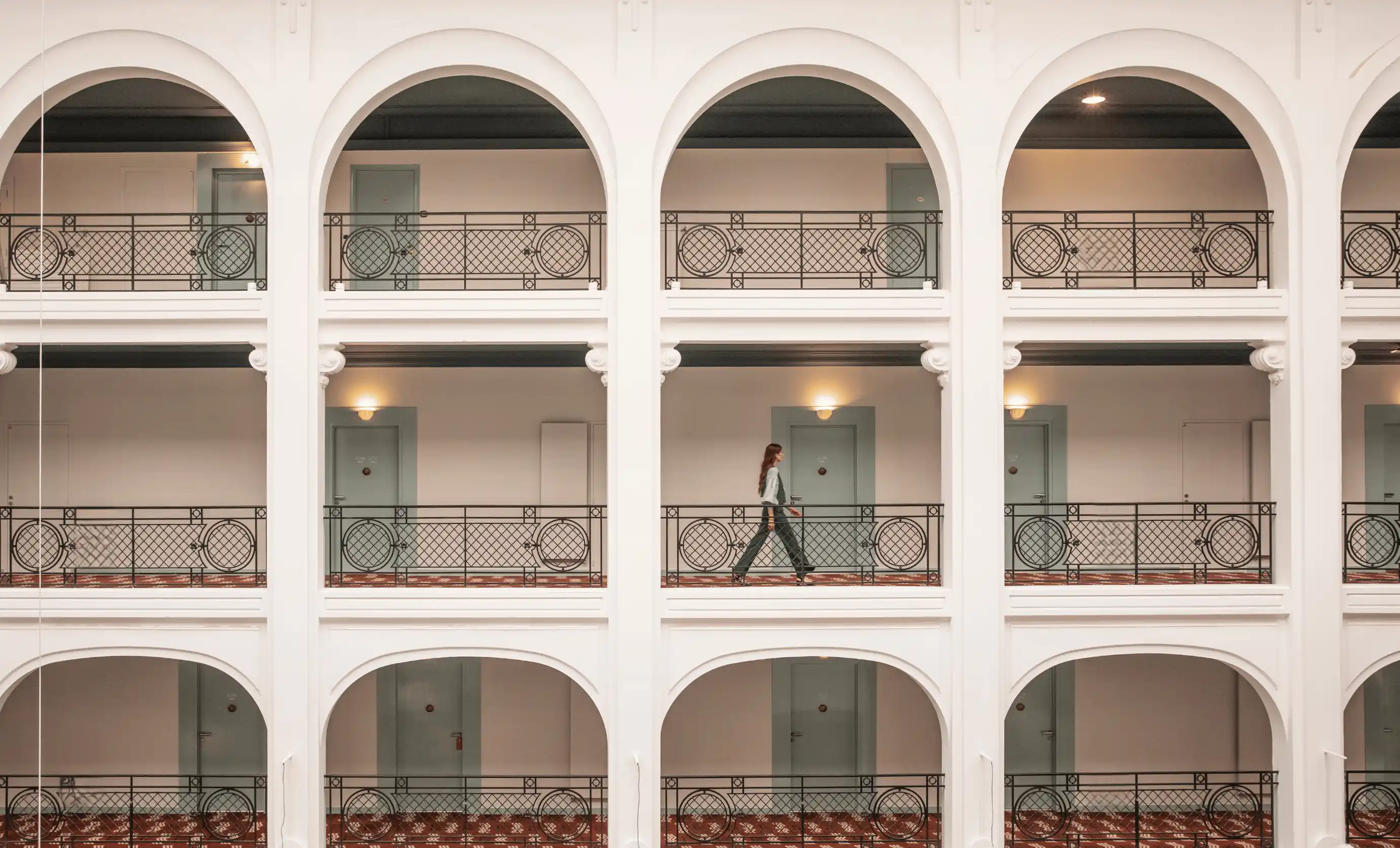 Woman walking along the second floor corridor of a hotel with white arches and decorative railings.