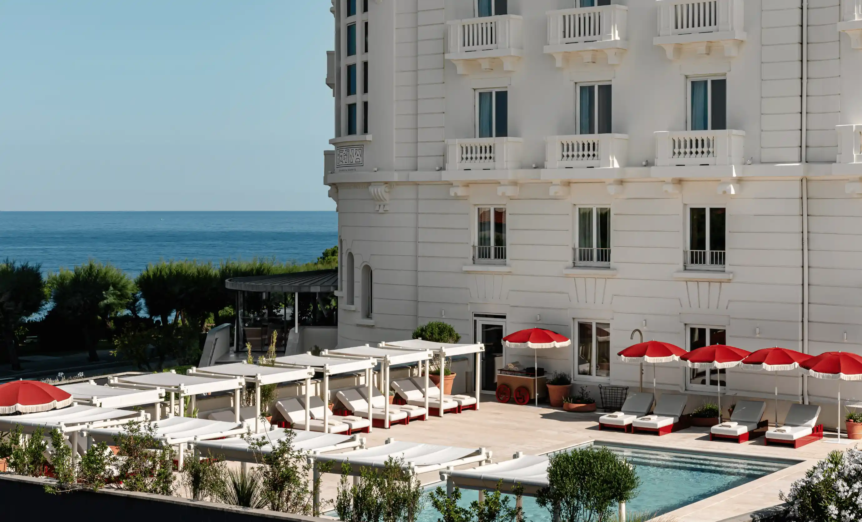 White hotel building with a poolside patio featuring white lounge chairs under red umbrellas and a view of the blue sea in the background.