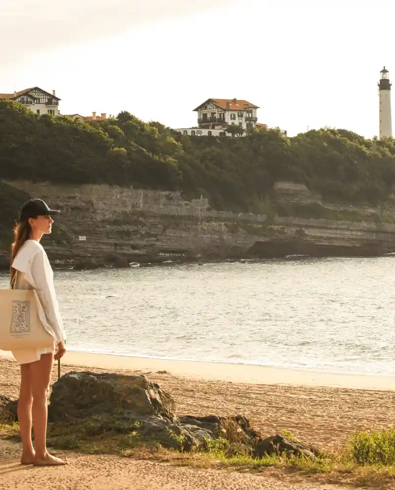 Woman standing barefoot on a beach path looking at the sea with coastal cliffs, houses, and a lighthouse in the background.
