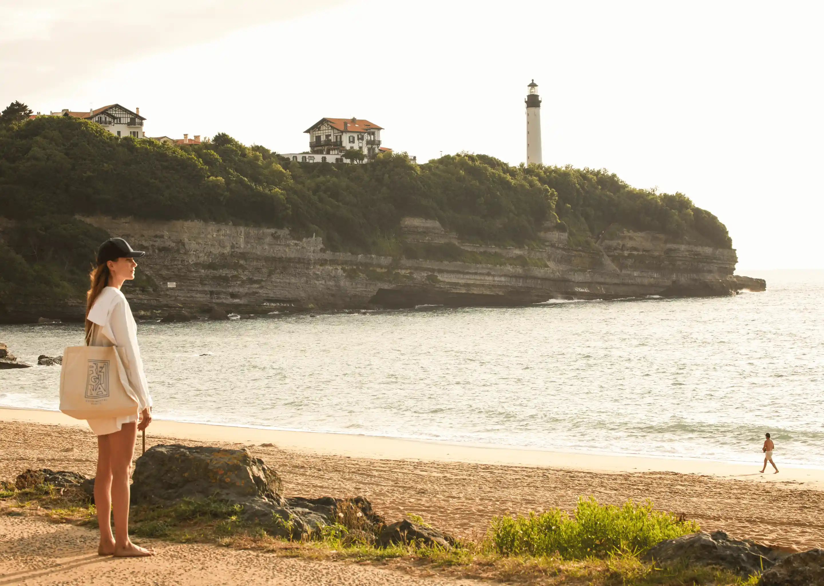 Woman in casual clothing and black cap standing on a rocky beach looking toward a cliff with houses and a lighthouse.