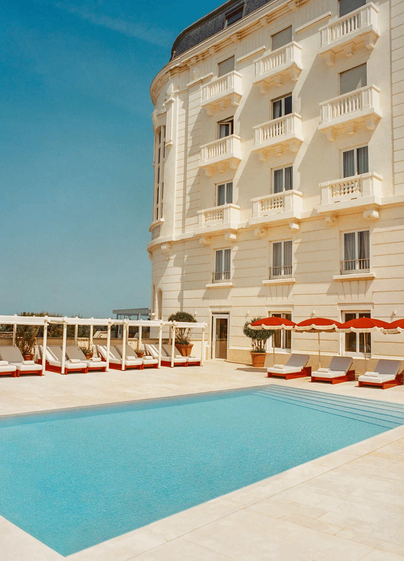 Outdoor swimming pool with lounge chairs and orange umbrellas beside a classic beige hotel building under a clear blue sky.