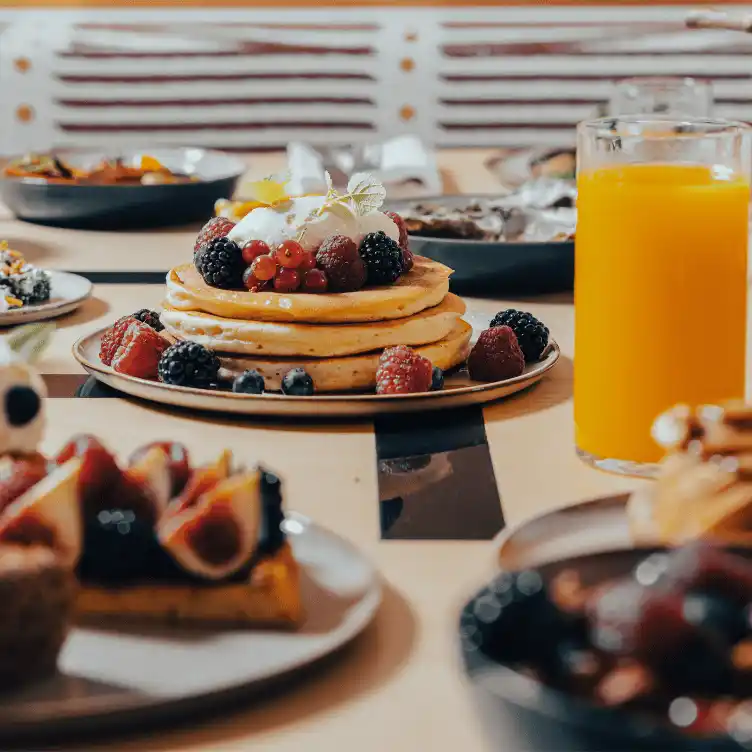 Stack of pancakes topped with whipped cream and mixed berries on a plate next to a glass of orange juice, with other breakfast dishes on the table.