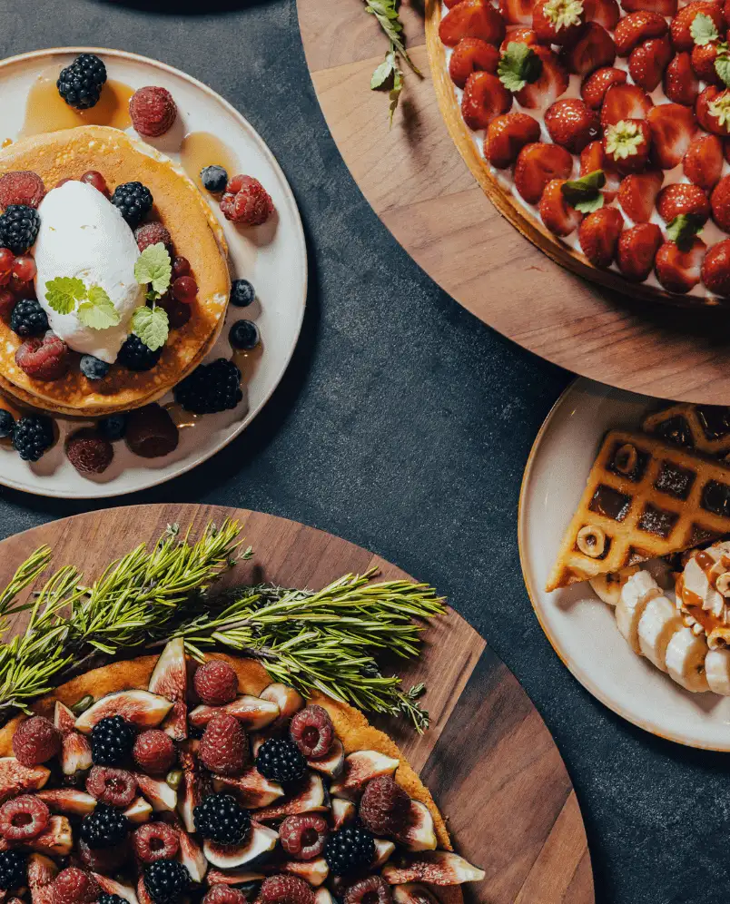 Assortment of brunch dishes including pancakes topped with berries and cream, a strawberry tart, a fruit and herb flatbread, and a waffle with bananas and syrup.