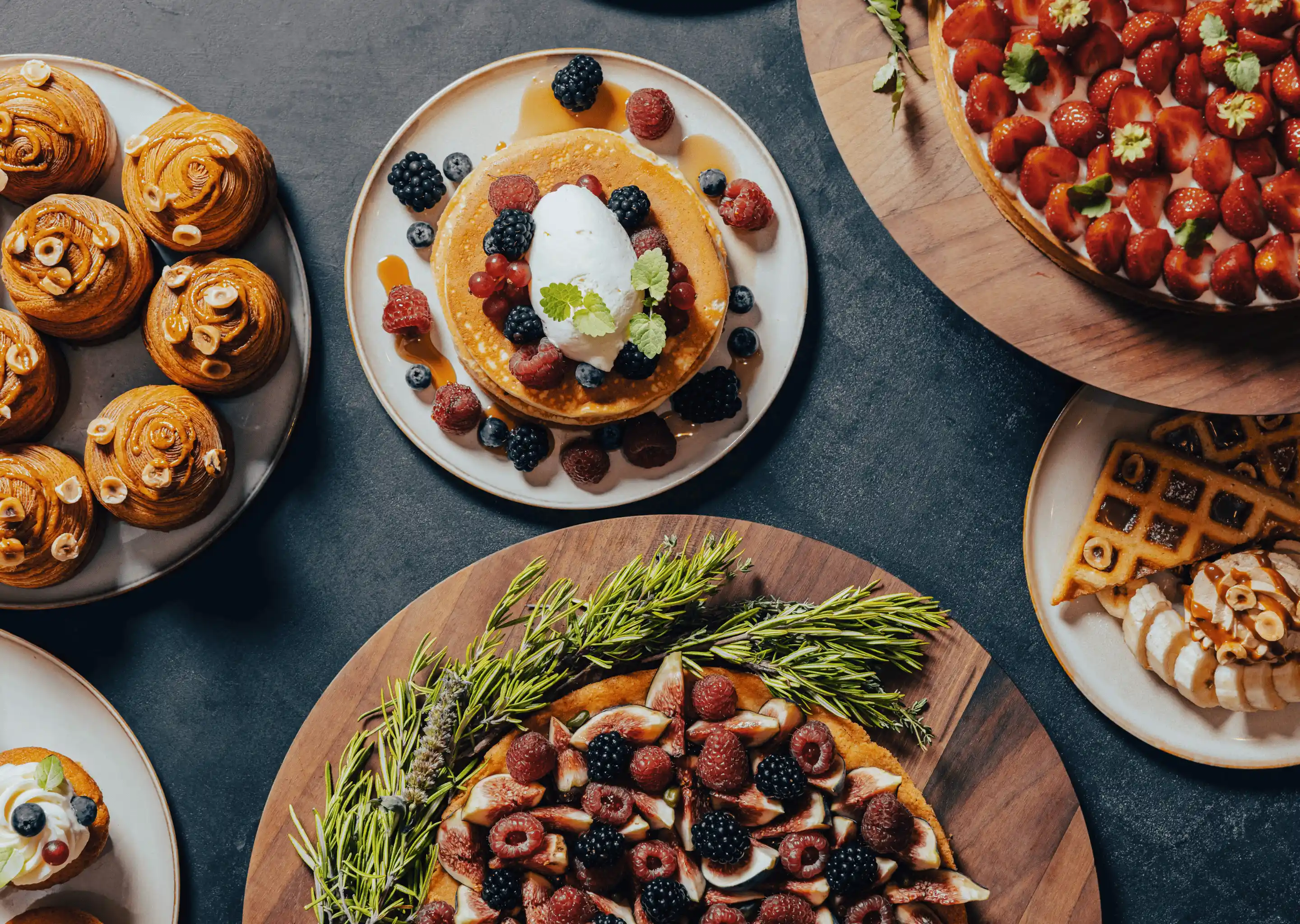 Assorted brunch dishes including pancakes with berries and cream, strawberry tart, chocolate swirled pastries, waffles with banana slices, and a fruit tart garnished with rosemary.