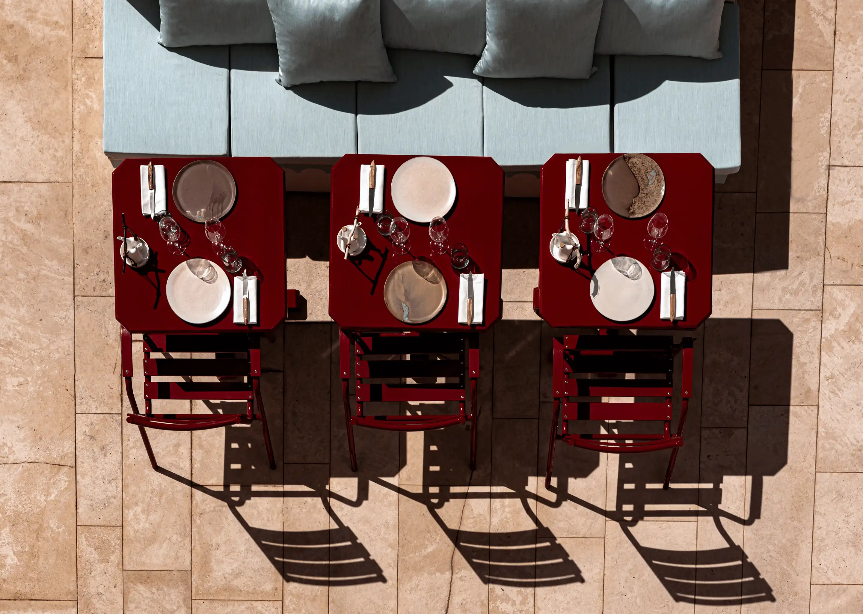 Aerial view of three red square tables with plates, napkins, and glasses, alongside a light blue cushioned bench on a tiled floor.