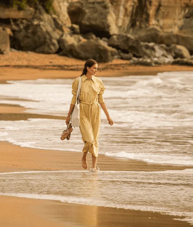 Woman in a long beige dress walking barefoot along a sandy beach with waves washing over her feet, holding sandals and a white shoulder bag, with rocky cliffs in the background.