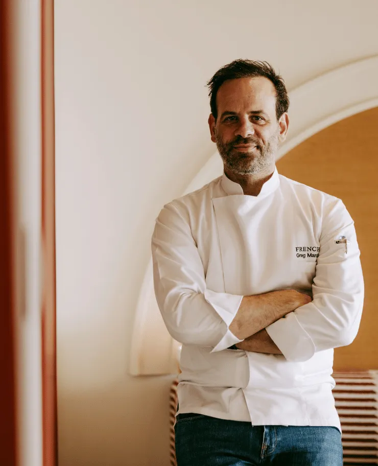 Man with short dark hair and beard wearing a white chef's jacket with folded arms, posing indoors.