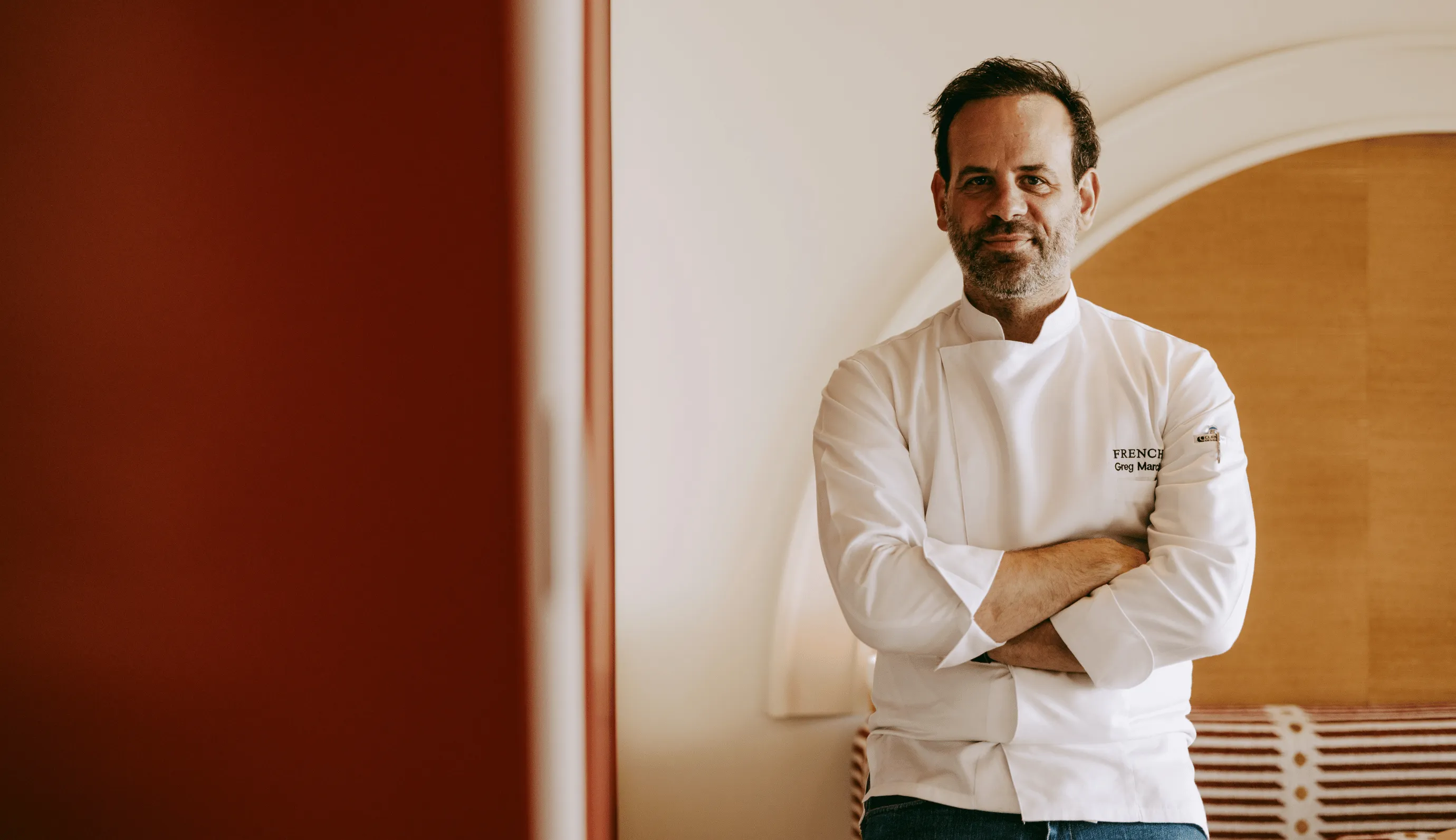 Male chef in white jacket stands with arms crossed against a light-colored wall with a red and white patterned bench.