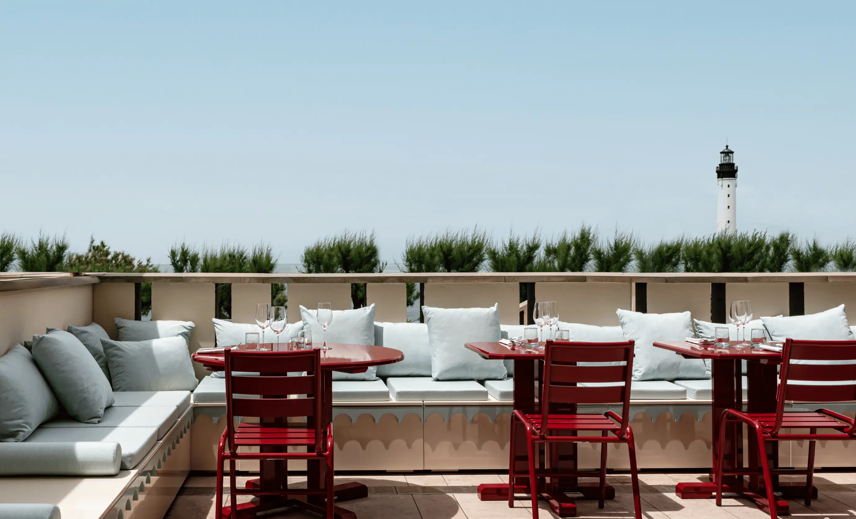 Outdoor terrace with red tables and chairs, light blue cushions on benches, greenery, and a white lighthouse in the distance under a clear sky.