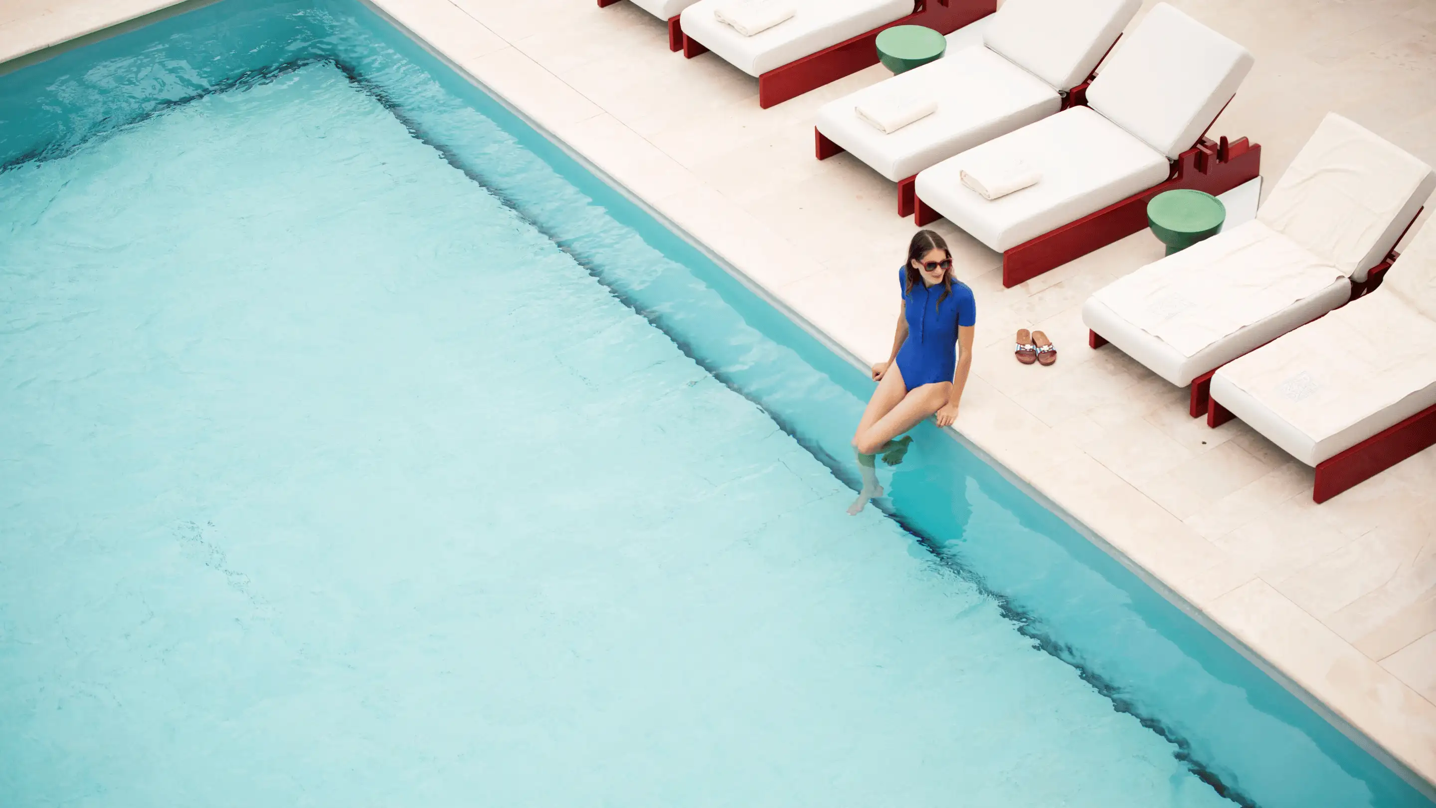 Woman in a blue swimsuit sitting at the edge of a pool with white lounge chairs and towels nearby.