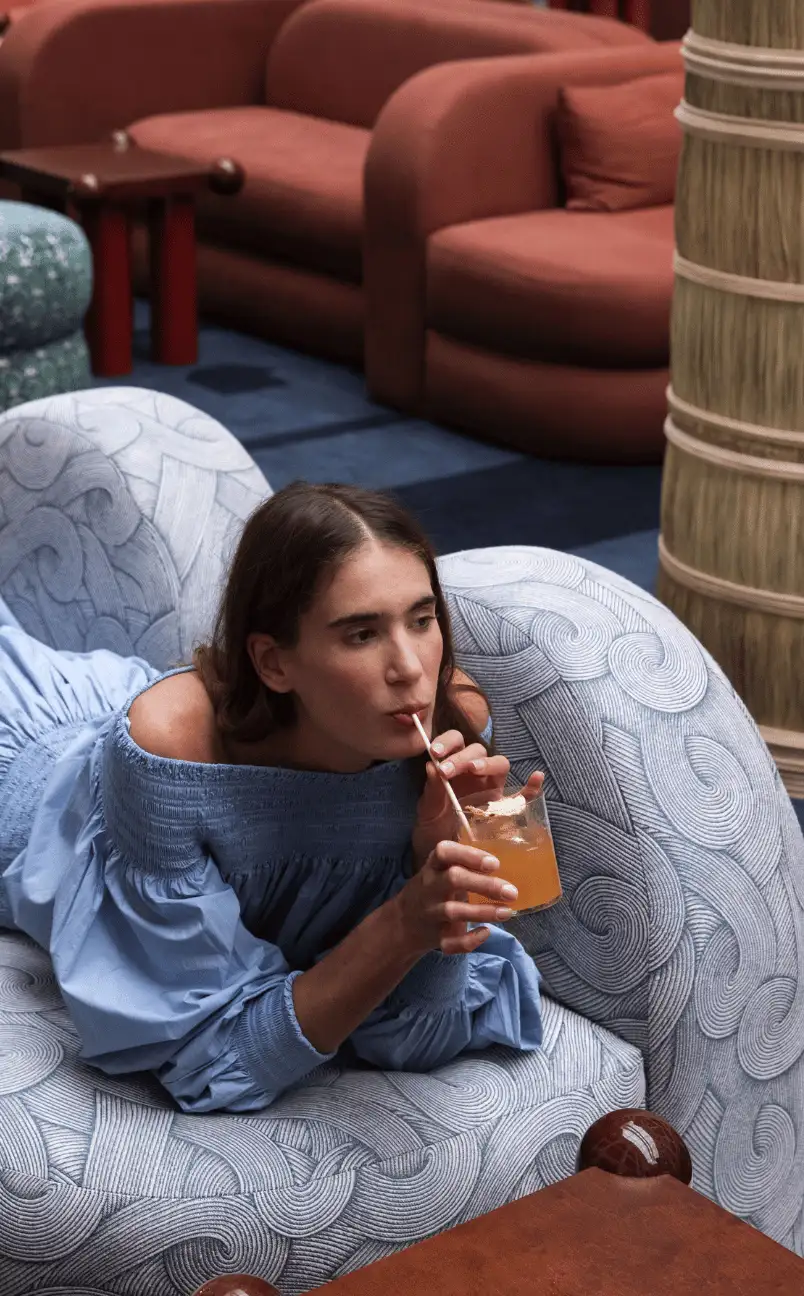 Woman in a blue off-shoulder dress lying on a patterned armchair, sipping an orange drink with a straw.