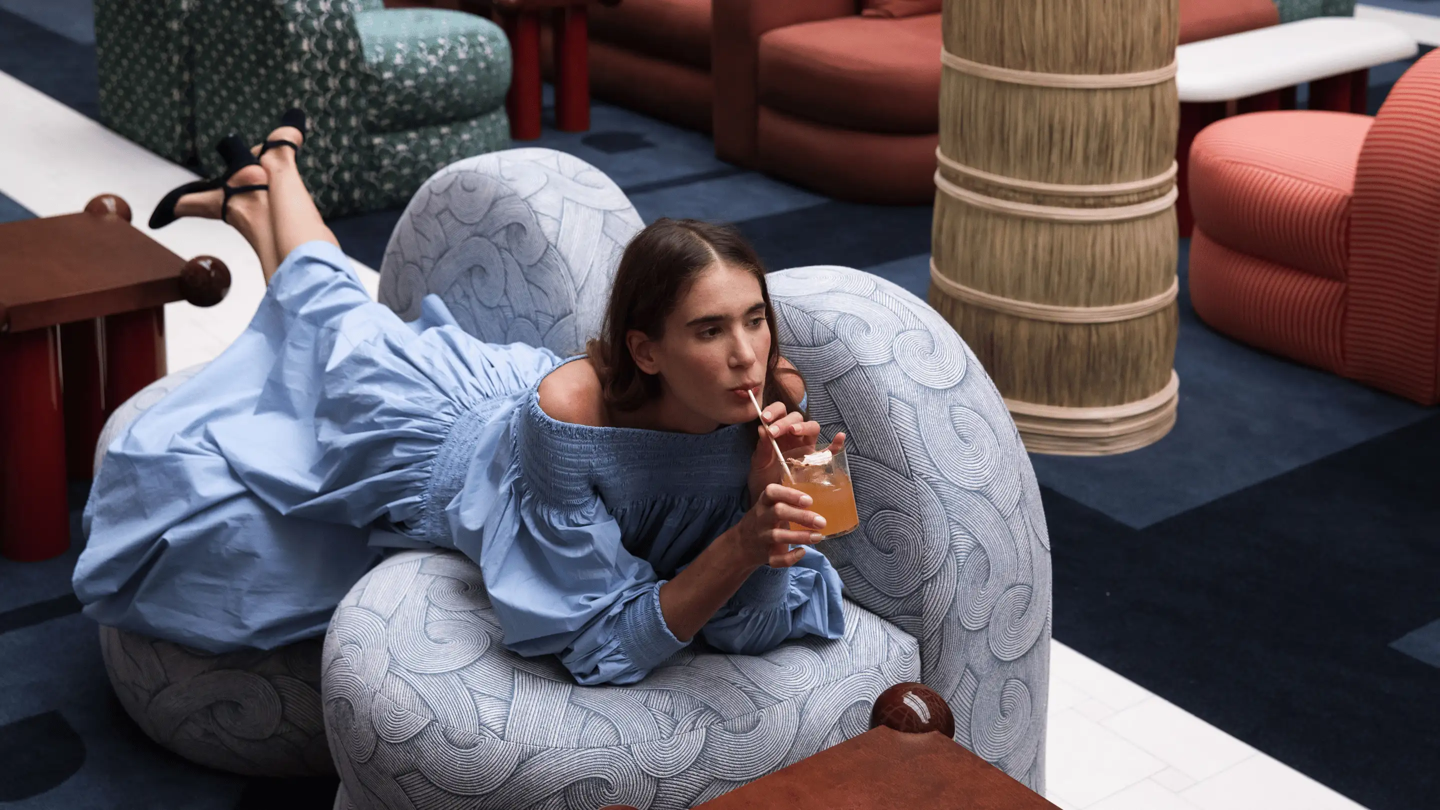 Woman in a blue dress lying on a patterned armchair drinking a cocktail with a straw in a stylish lounge.