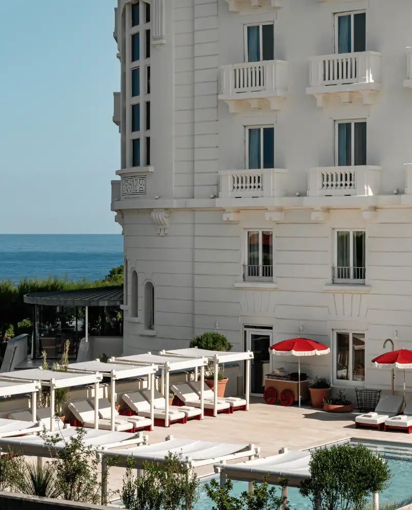 Outdoor pool area with white lounge chairs and red umbrellas beside a white multi-story building overlooking the ocean.