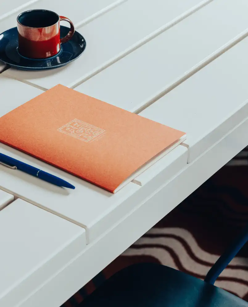 Orange notebook, blue pen, and red cup with saucer on a white slatted table with a black chair and a patterned rug underneath.