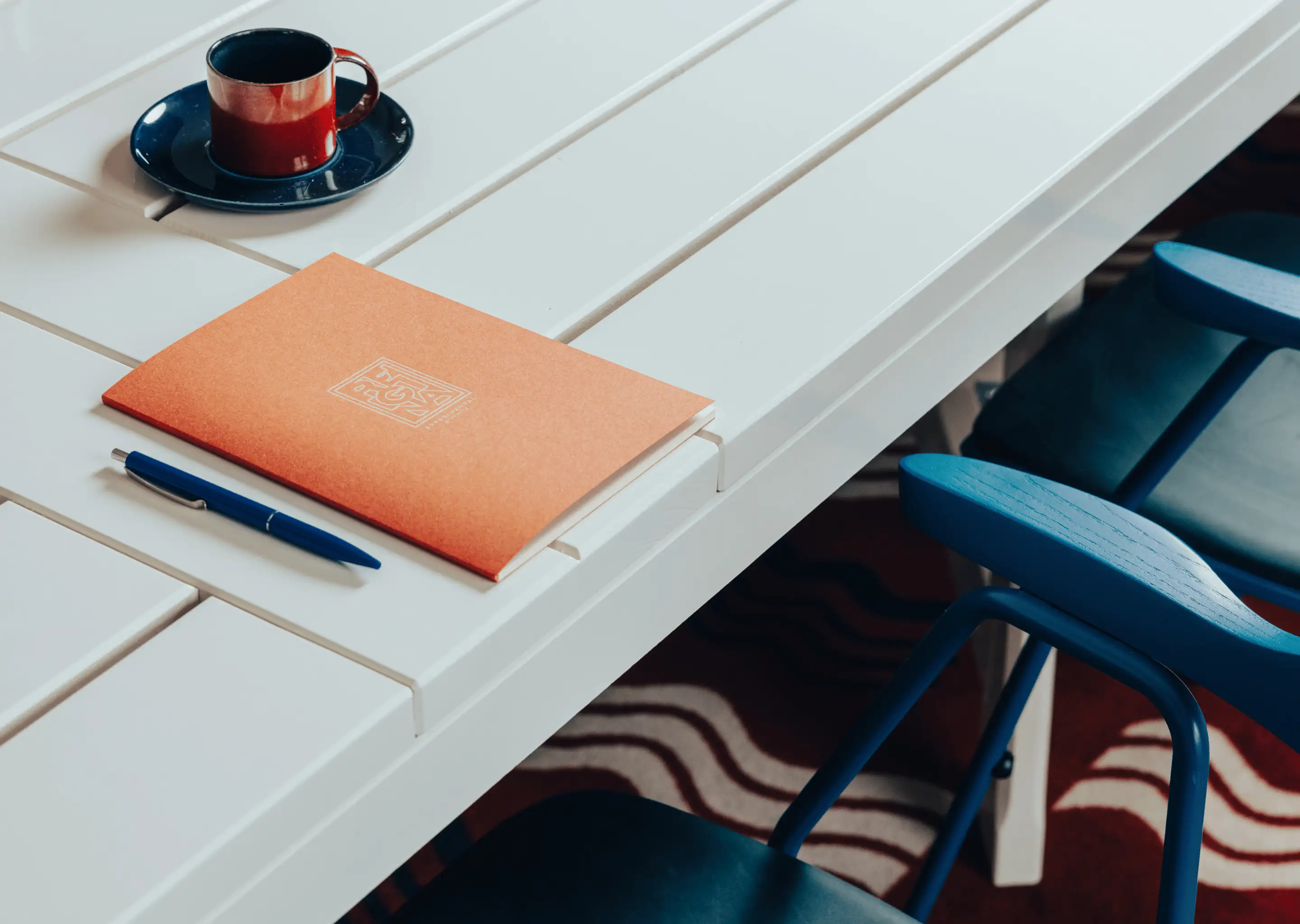 White slatted table with an orange notebook, blue pen, and a brown cup with a black saucer; blue chairs are beside the table.