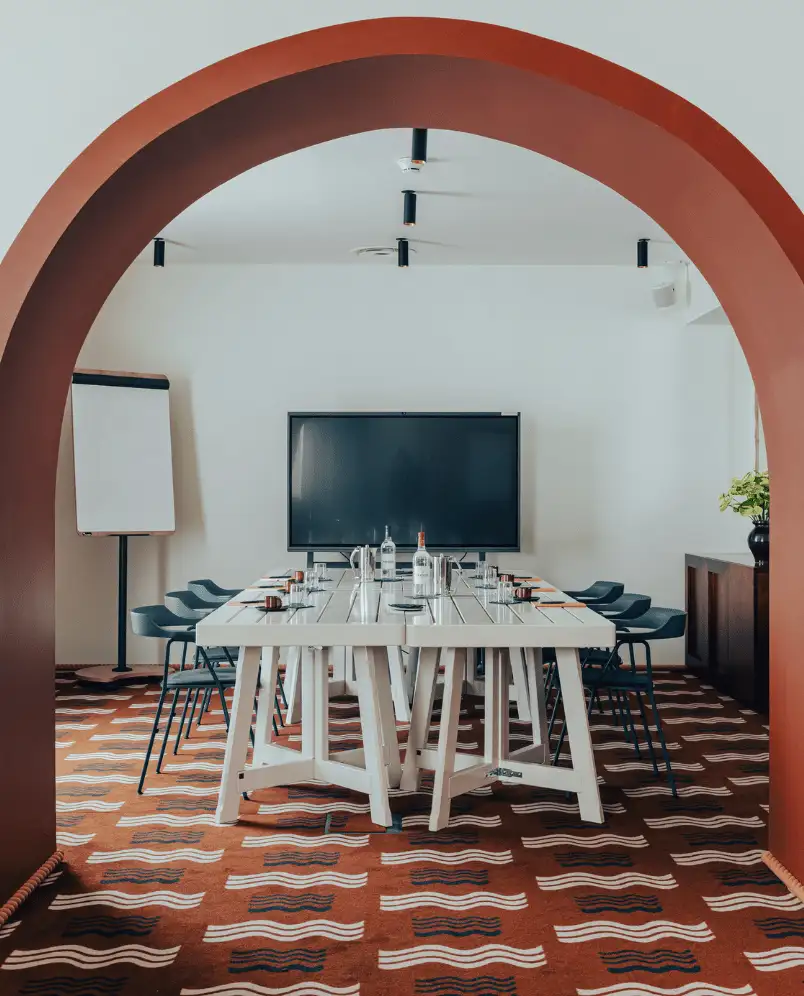 Modern conference room with a white rectangular table, black chairs, a large flat screen TV, and a white flip chart, viewed through a large terracotta archway.