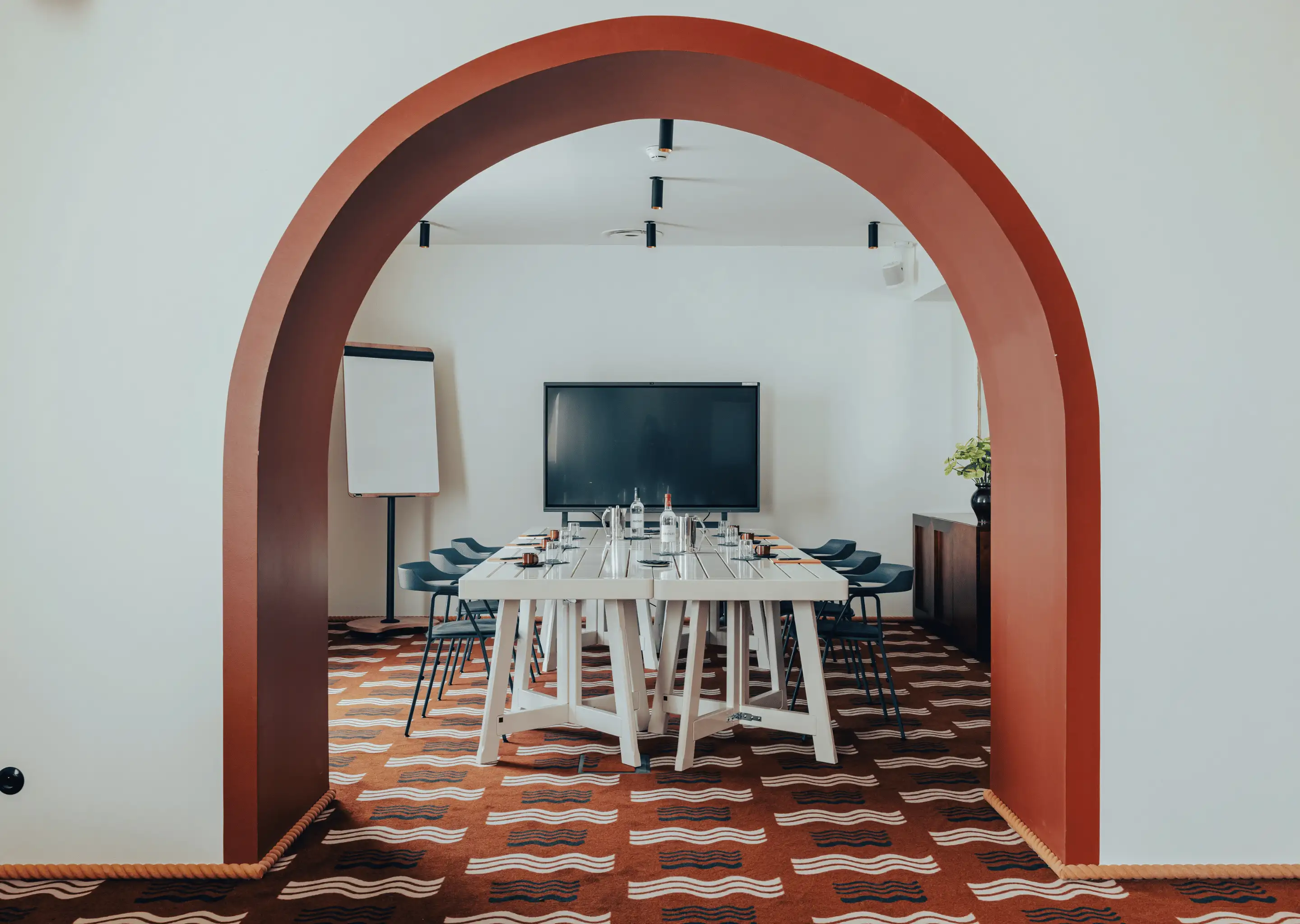Conference room with a white table, black chairs, a large screen, a flip chart, and a patterned carpet visible through a red arched doorway.