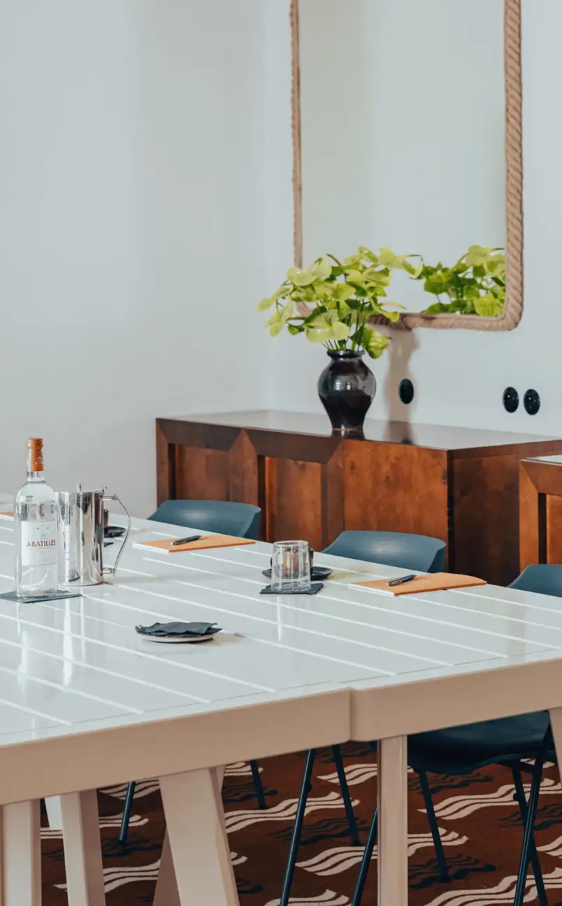 Meeting room with white table, blue chairs, notebooks, glassware, and a wooden cabinet with a black vase of green flowers under a large mirror.