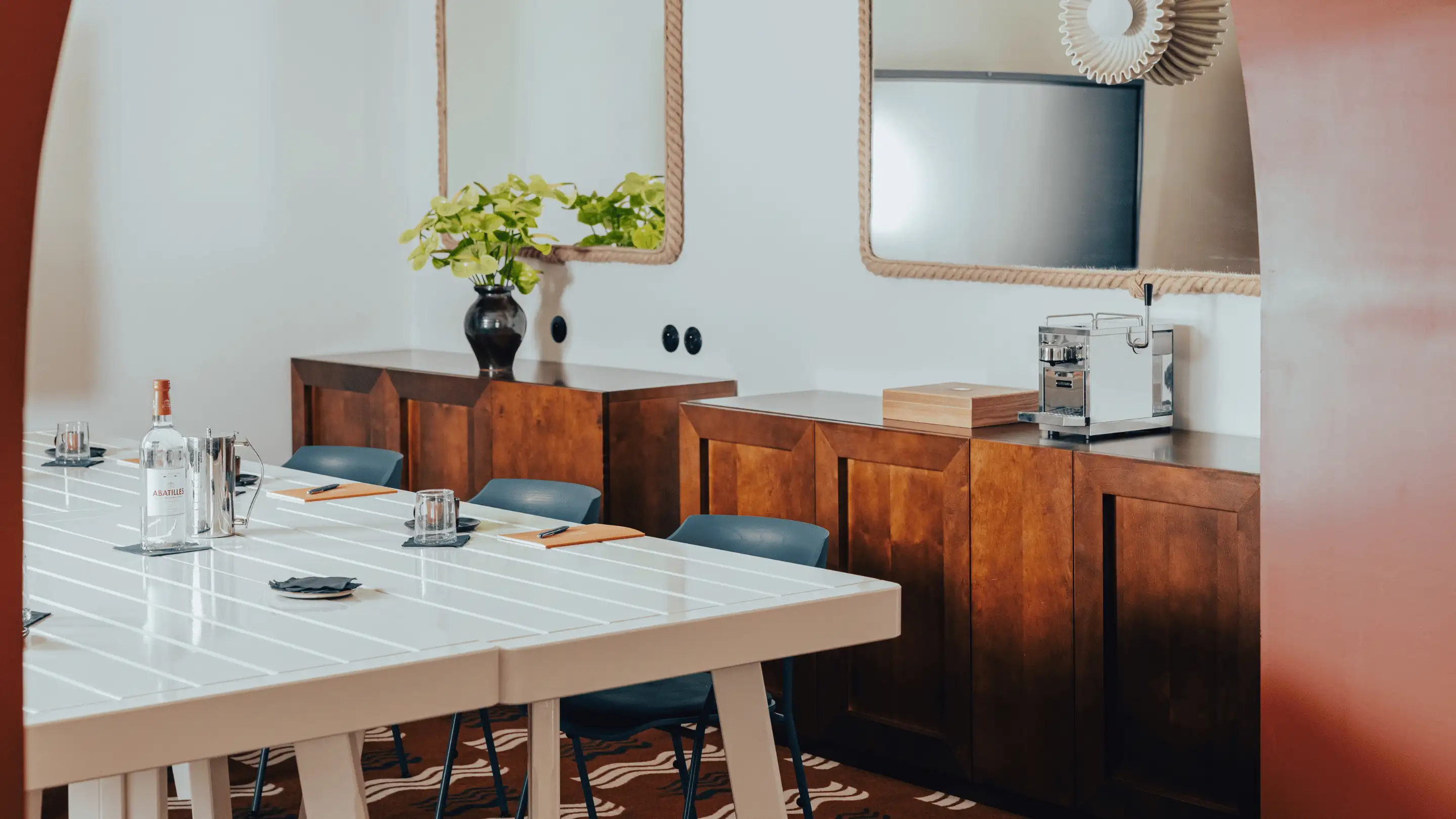 Modern meeting room with a white table, blue chairs, wooden cabinets, a water bottle, glasses, and a coffee machine.