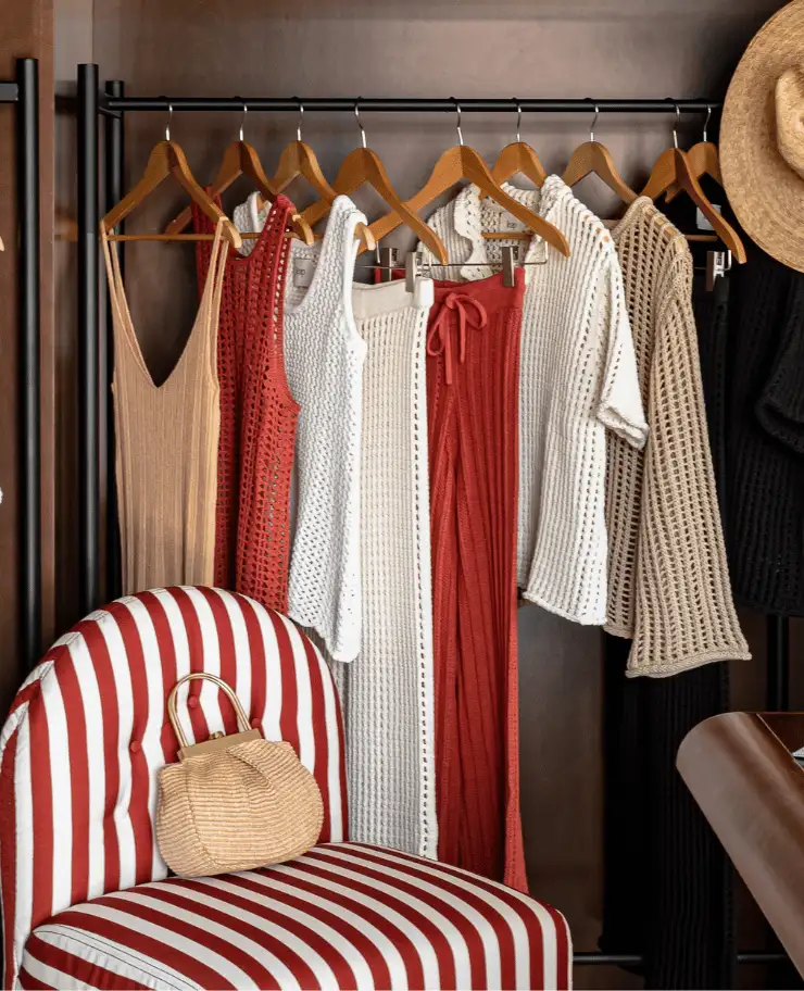 Boutique closet with hanging knitted clothing in beige, white, rust, and black colors, and a striped red and white chair holding a woven handbag.
