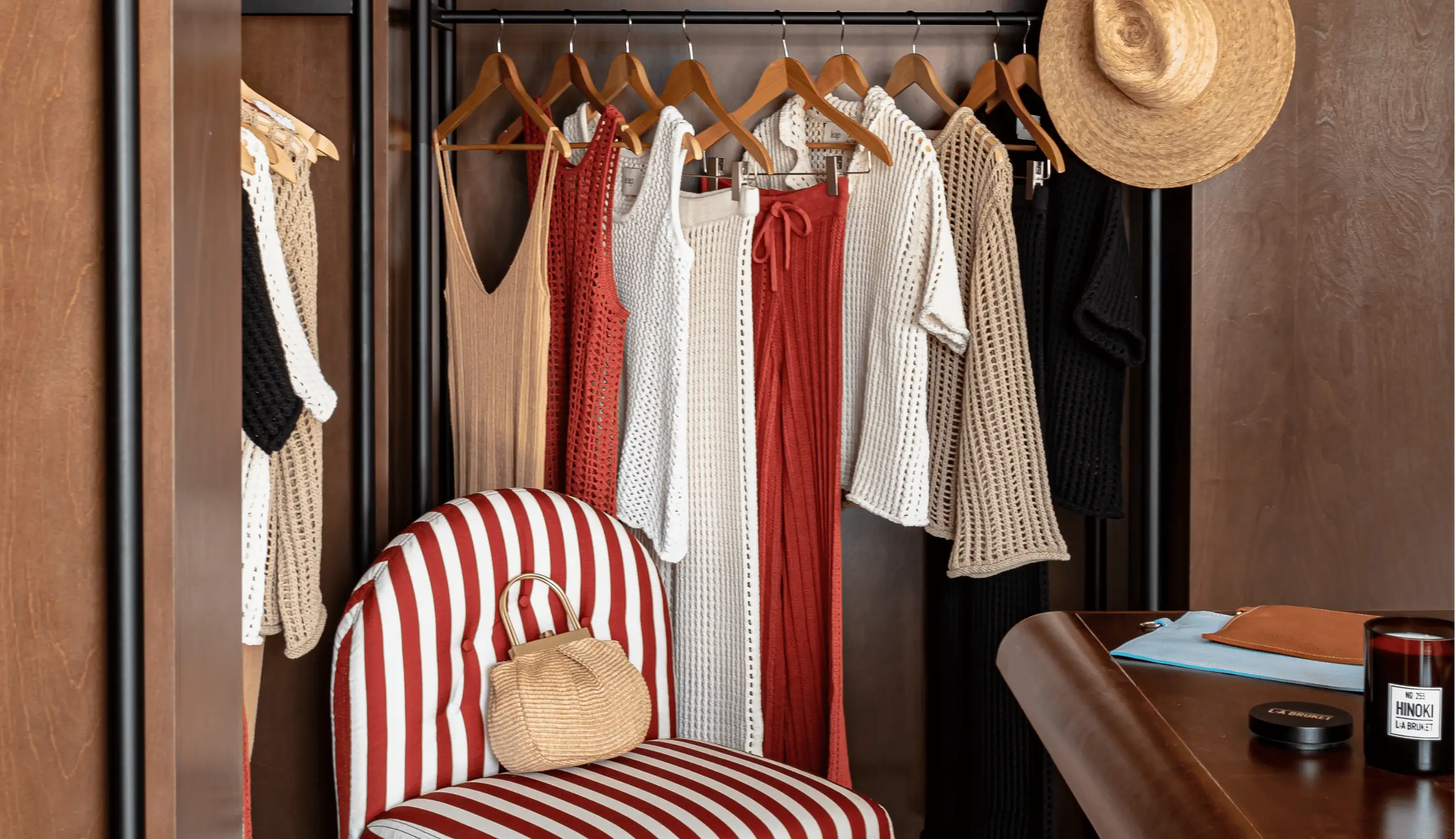 Boutique closet with crocheted summer dresses and tops in neutral and red tones hanging on wooden hangers, a striped chair with a woven handbag, and a straw hat hanging on the side.