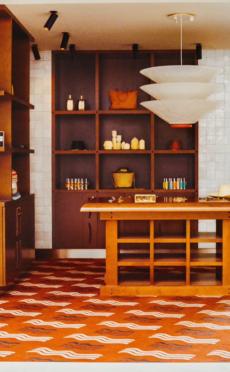 Interior of a boutique store with wooden shelves displaying bags, candles, and small bottles, an orange patterned floor, and a hanging white lamp.