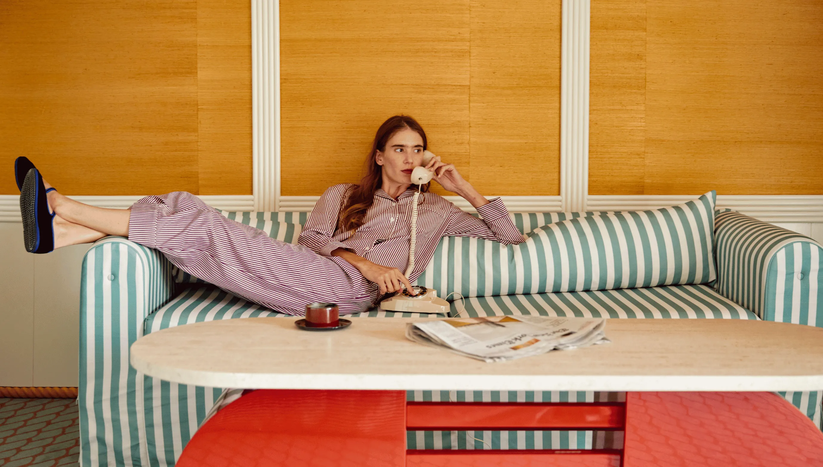 Woman in striped pajamas lying on a striped couch, talking on a cream rotary phone with a coffee cup and newspaper on the table in front.
