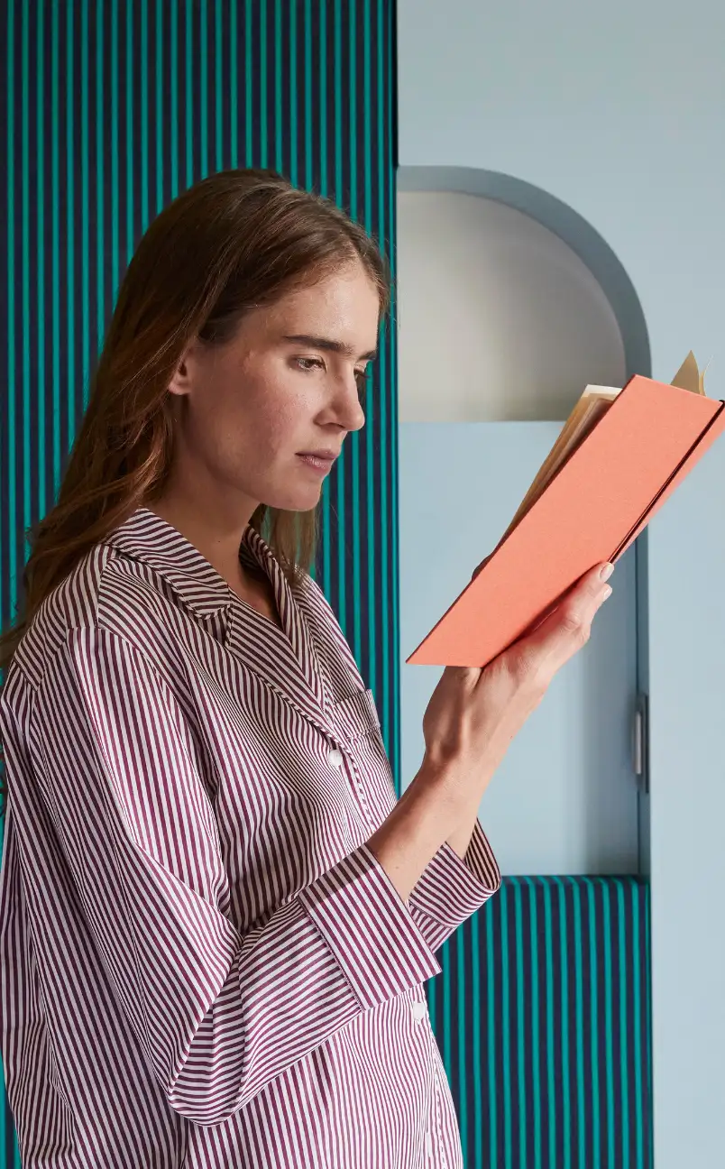 Woman in striped shirt reading a coral-colored book against a teal and blue patterned background.