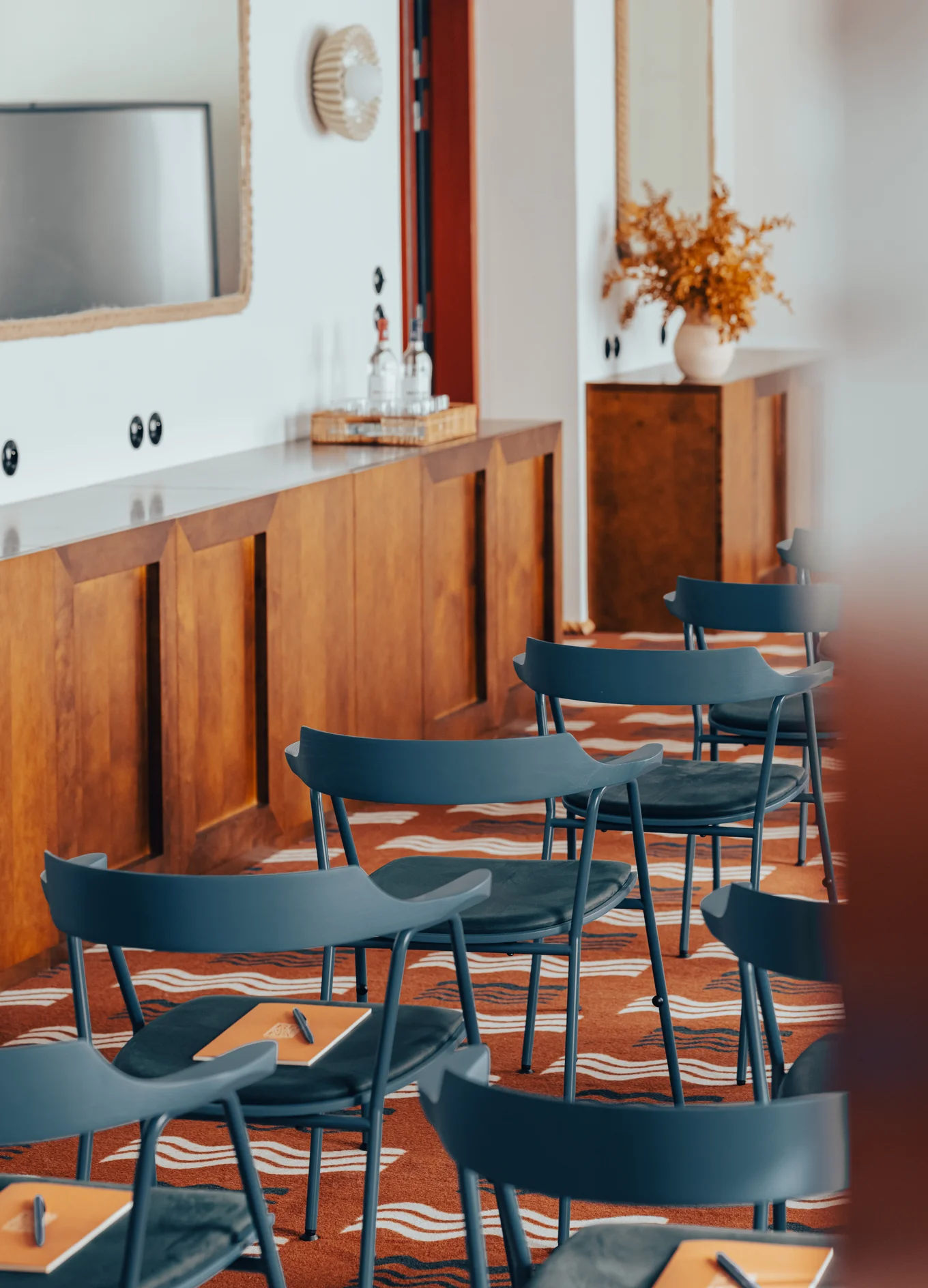 A room set up with blue chairs arranged in rows on a patterned carpet, wooden cabinets along the wall, and a vase with dried flowers on a cabinet in the background.