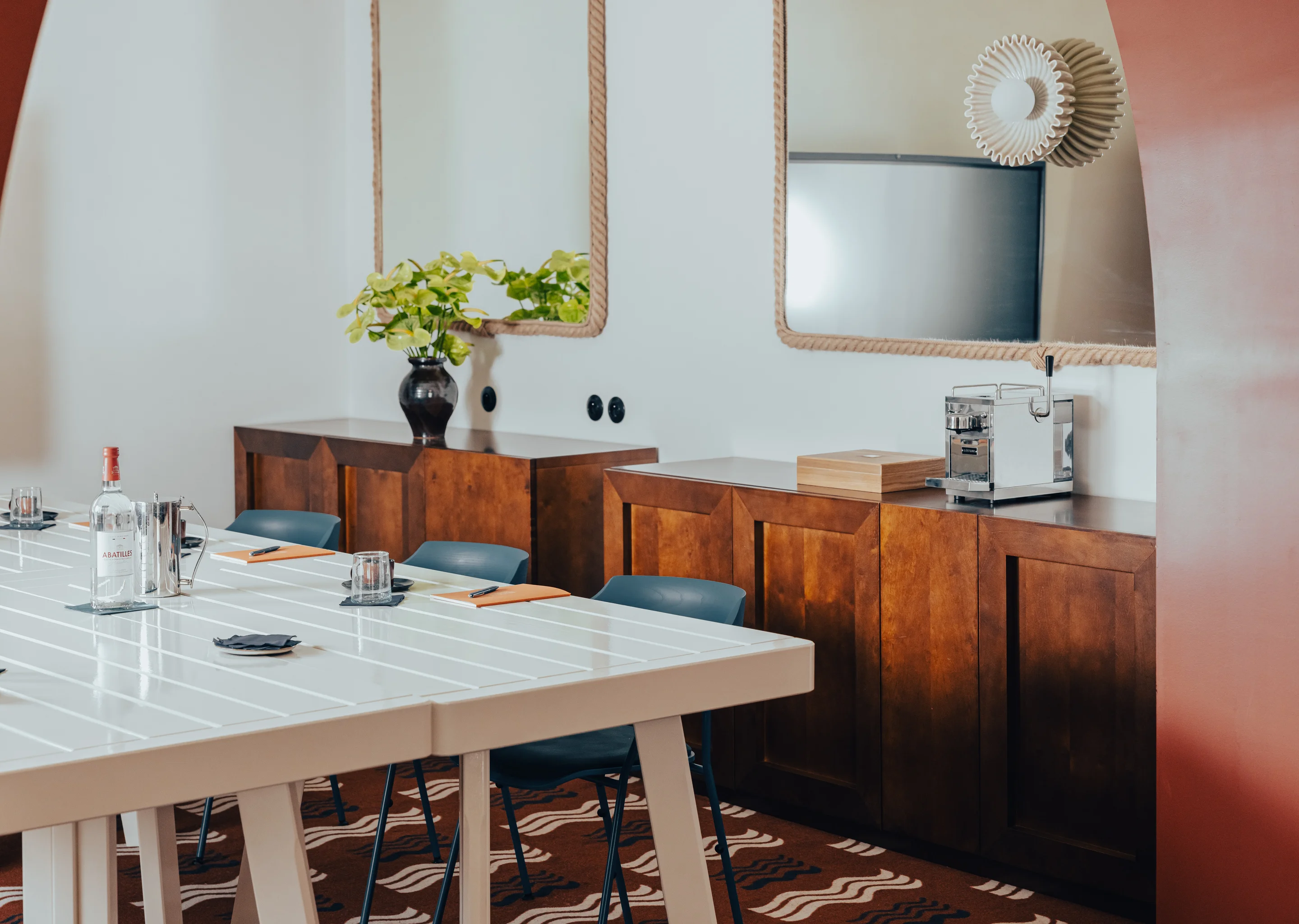 Modern meeting room with white table, blue chairs, wooden cabinets, two rope-framed mirrors, and a plant in a black vase.