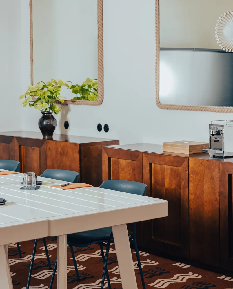 Modern meeting room with white table, blue chairs, wooden cabinets, two rope-framed mirrors, a black vase with green leaves, and a coffee machine.