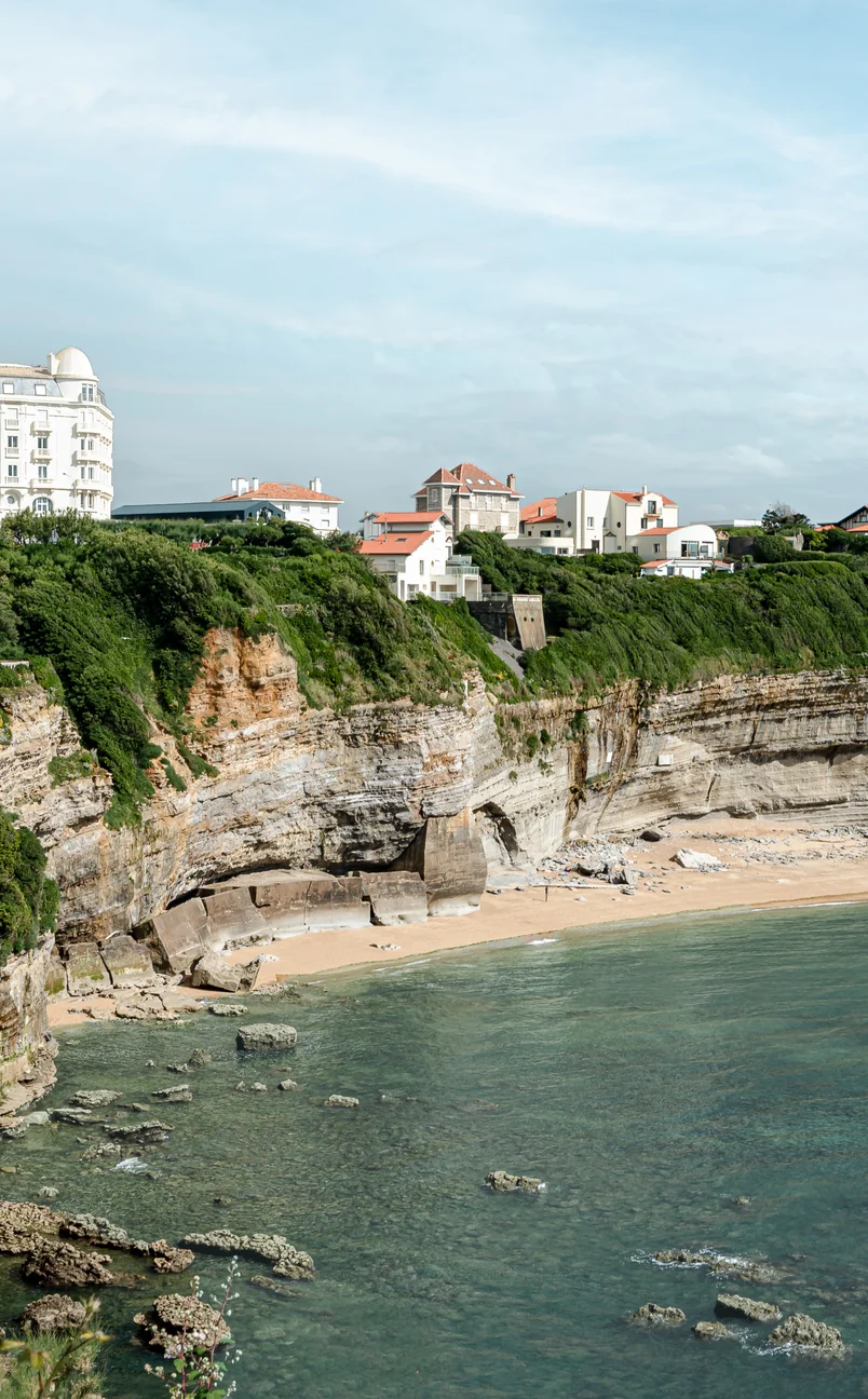 Cliffside beachfront with rocky shore and houses with red roofs atop green cliffs under a cloudy sky.