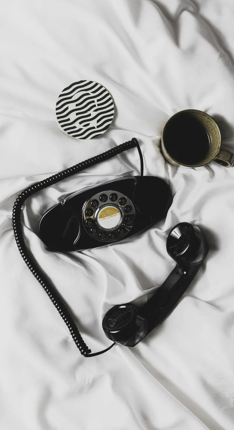 Vintage black rotary dial telephone with handset off the hook, a coffee cup, and a black-and-white striped coaster on white fabric.