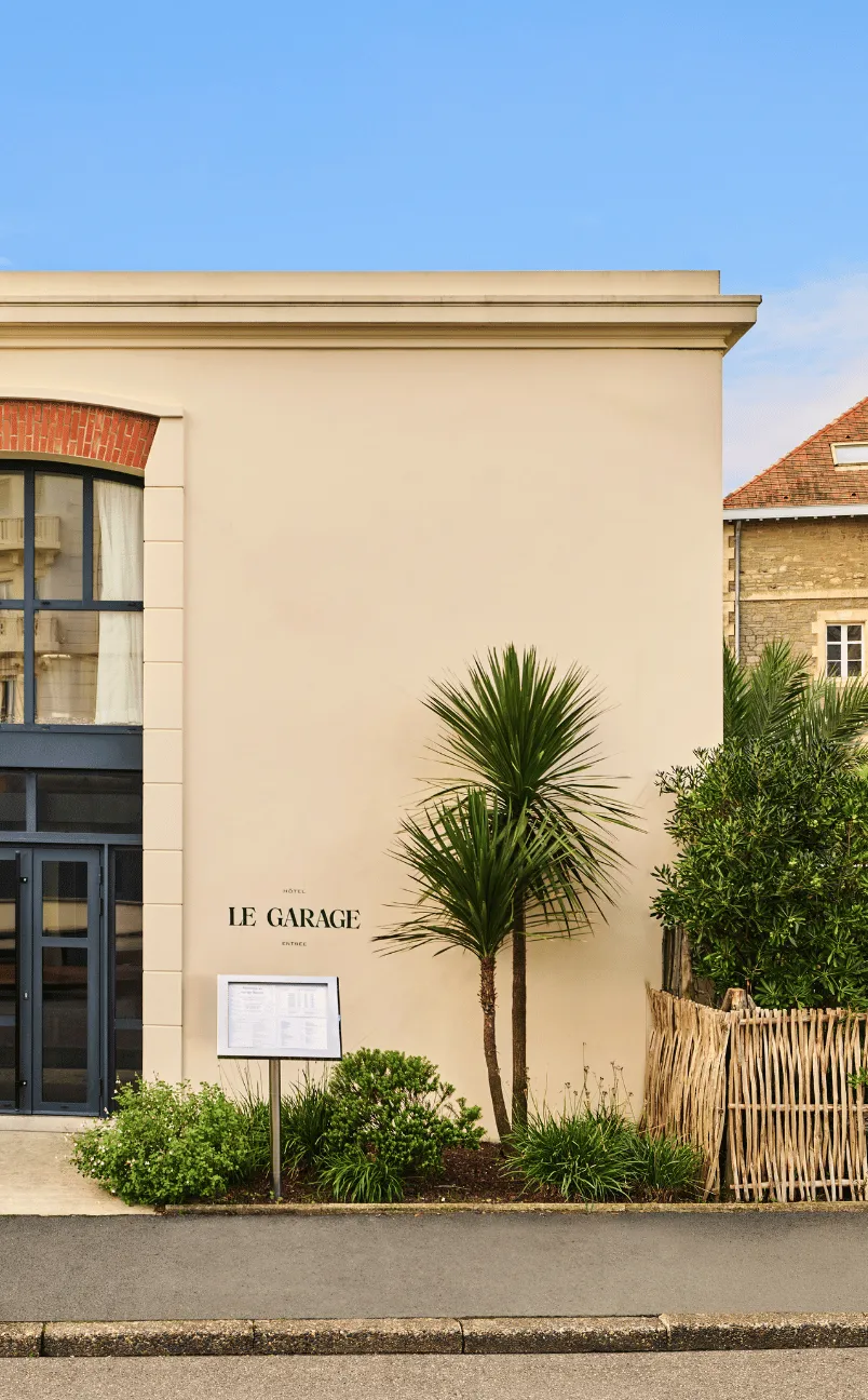 The exterior entrance of Hotel Le Garage with a beige wall, glass door, tropical plants, and a menu display stand.