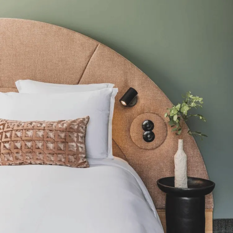 Bed with white linens, beige textured semicircular headboard, decorative pillow, black spotlight, and round side table with a white vase holding green leaves against a muted green wall.