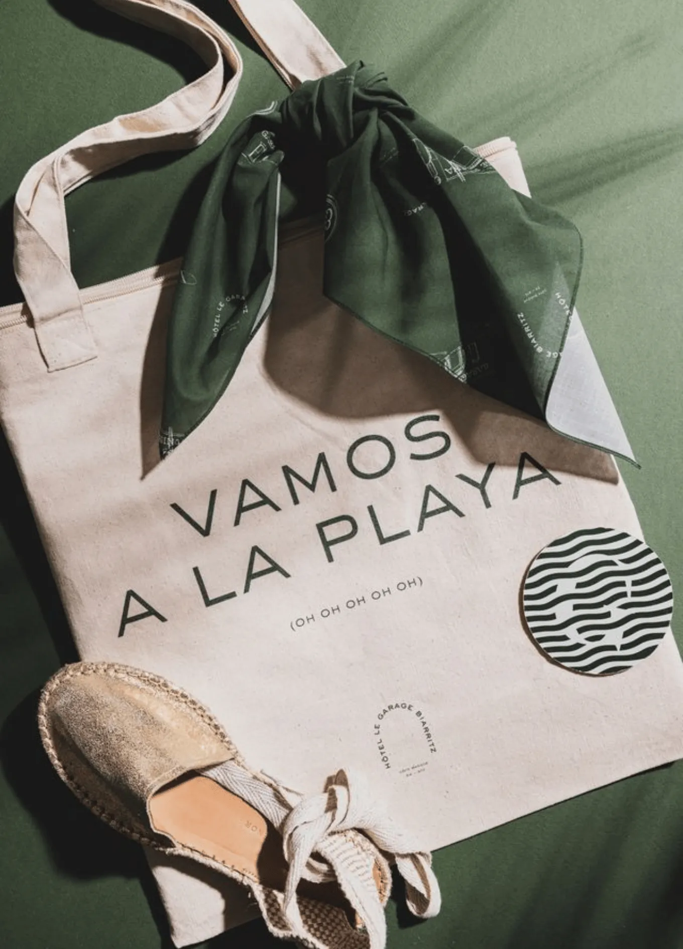 Beige tote bag with text 'VAMOS A LA PLAYA' and a green scarf tied to the handle, placed on a green surface with a pair of beige espadrilles and a black and white patterned round coaster.