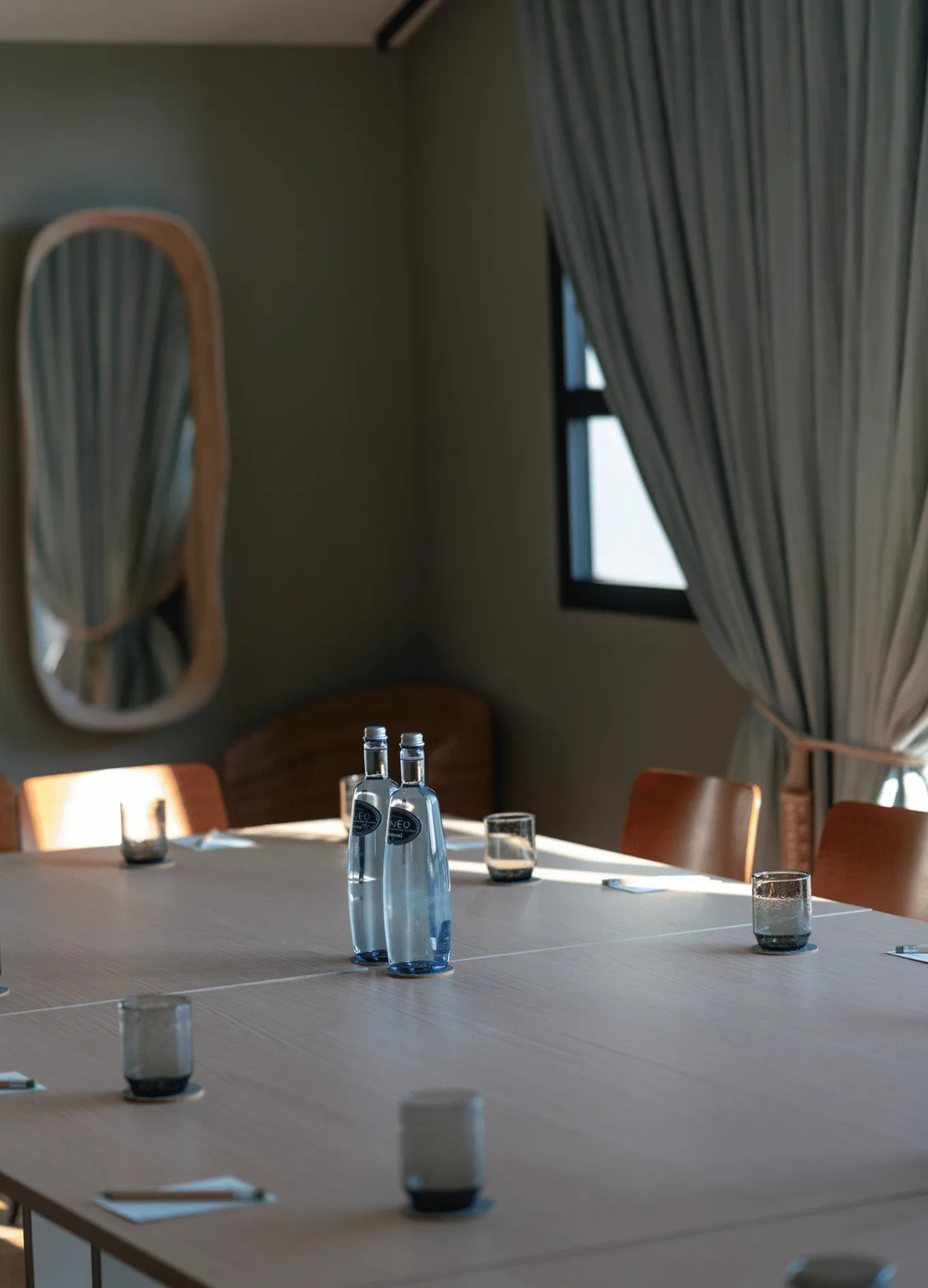 Modern conference room with wooden table, brown chairs, two water bottles, empty glasses, and natural light from a window with curtains.
