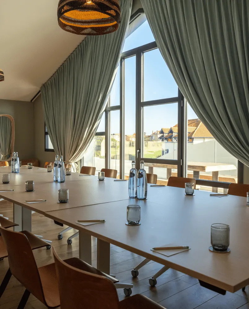 Modern conference room with long white tables, brown chairs, water bottles, glasses, and large windows with green curtains.