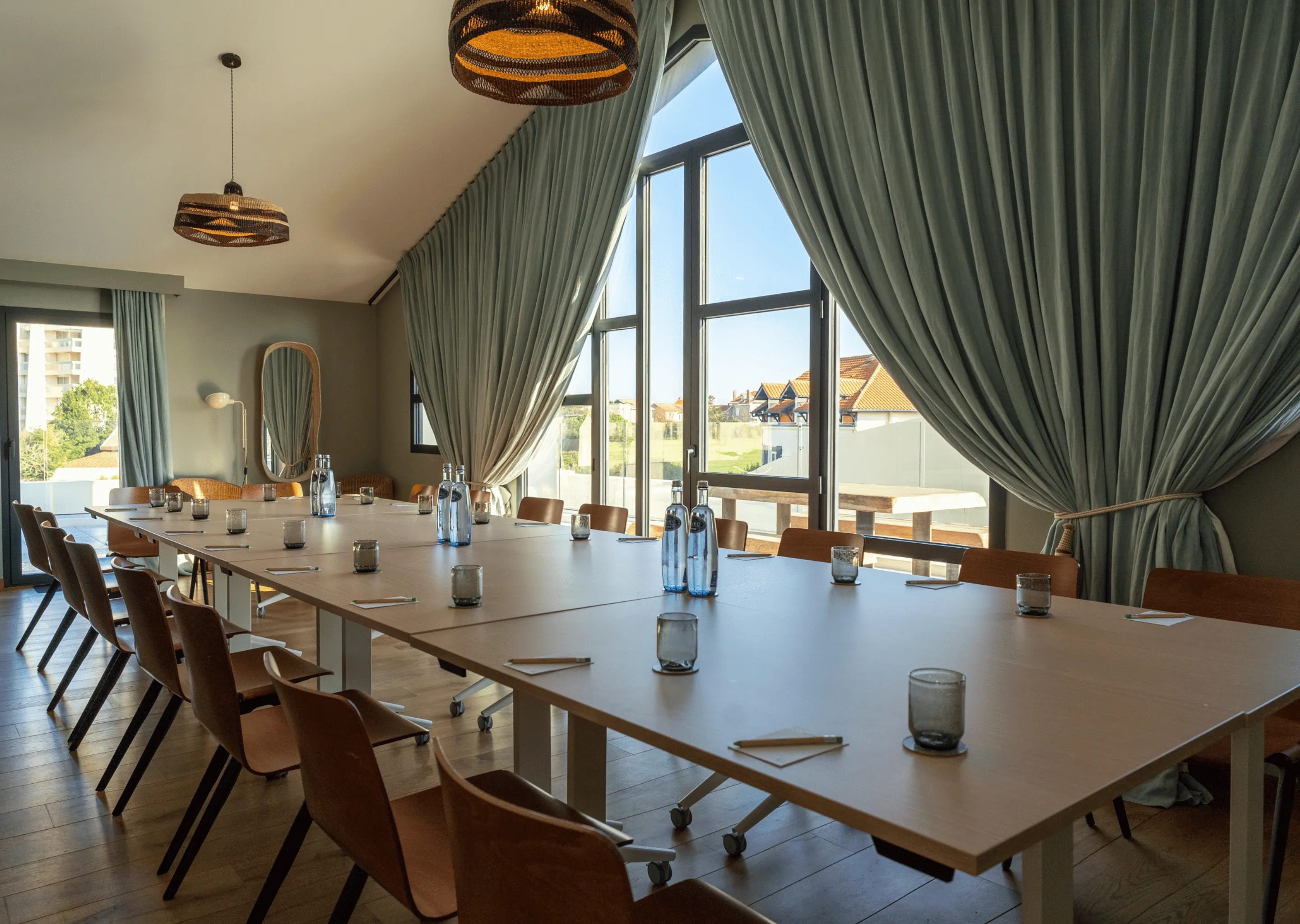 Conference room with a large rectangular table surrounded by wooden chairs, set with glasses, notepads, and water bottles, illuminated by natural light from tall windows with curtains.