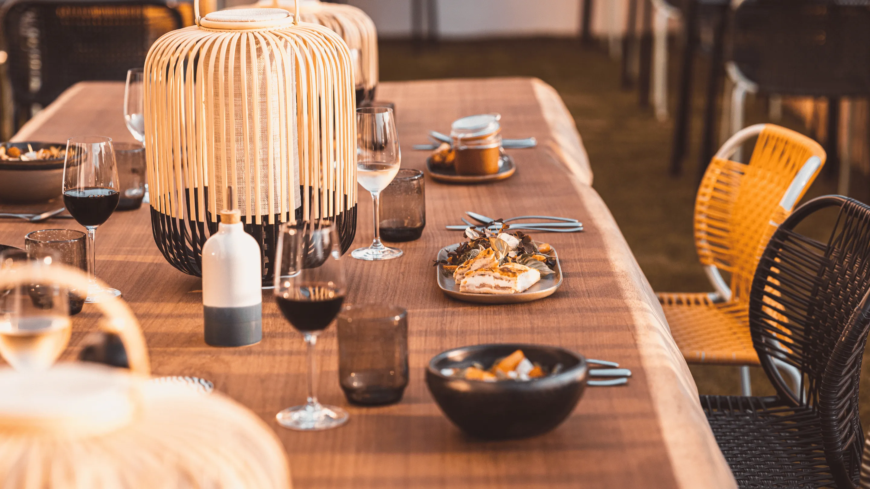 Long wooden dining table set with wine glasses, decorative lanterns, plates with food, and woven chairs in warm lighting.