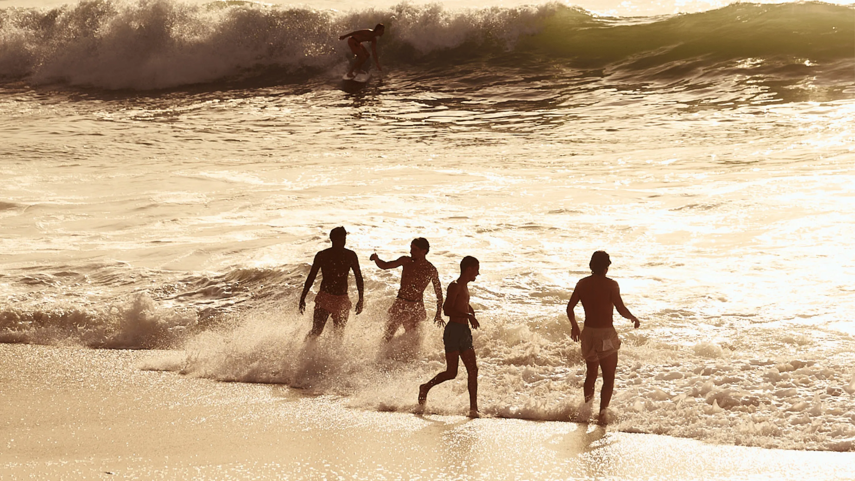 Four boys playing and splashing in shallow ocean water near the shore at sunset with a surfer riding a wave in the background.