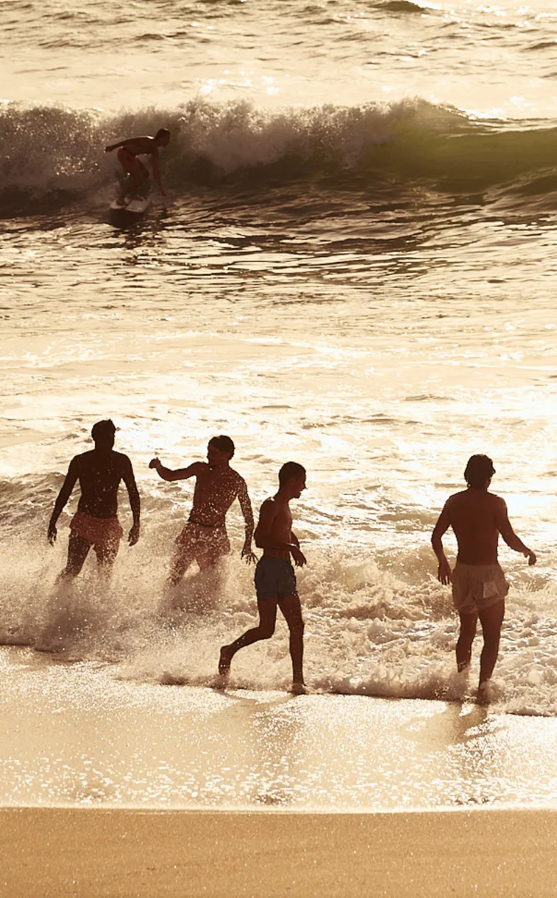 Four young men playing at the edge of the ocean shore while another person surfs a wave in the background during sunset.